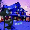 Two styles of snowflake projection lights with ground stake and wall mount base, projecting multi-color snowflake patterns on snow covered farmhouse at dusk, winter Christmas outdoor decor product comparison display